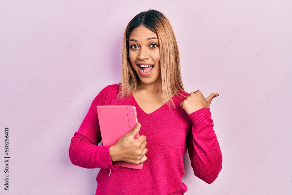 Beautiful hispanic woman holding book pointing thumb up to the side smiling happy with open mouth Beautiful hispanic woman holding book pointing thumb up to the side smiling happy with open mouth