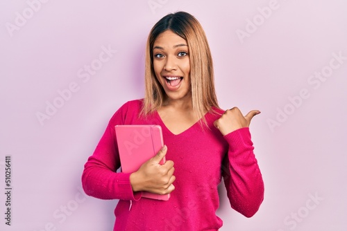 Beautiful hispanic woman holding book pointing thumb up to the side smiling happy with open mouth