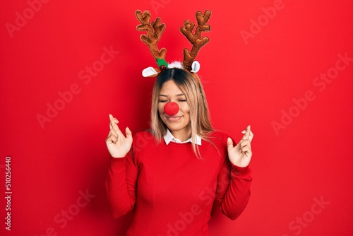 Beautiful hispanic woman wearing deer christmas hat and red nose gesturing finger crossed smiling with hope and eyes closed. luck and superstitious concept.