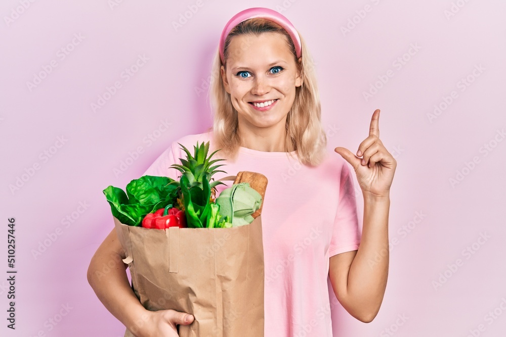 Beautiful caucasian blonde woman holding paper bag with groceries smiling happy pointing with hand and finger to the side Beautiful caucasian blonde woman holding paper bag with groceries smiling happy pointing with hand and finger to the side