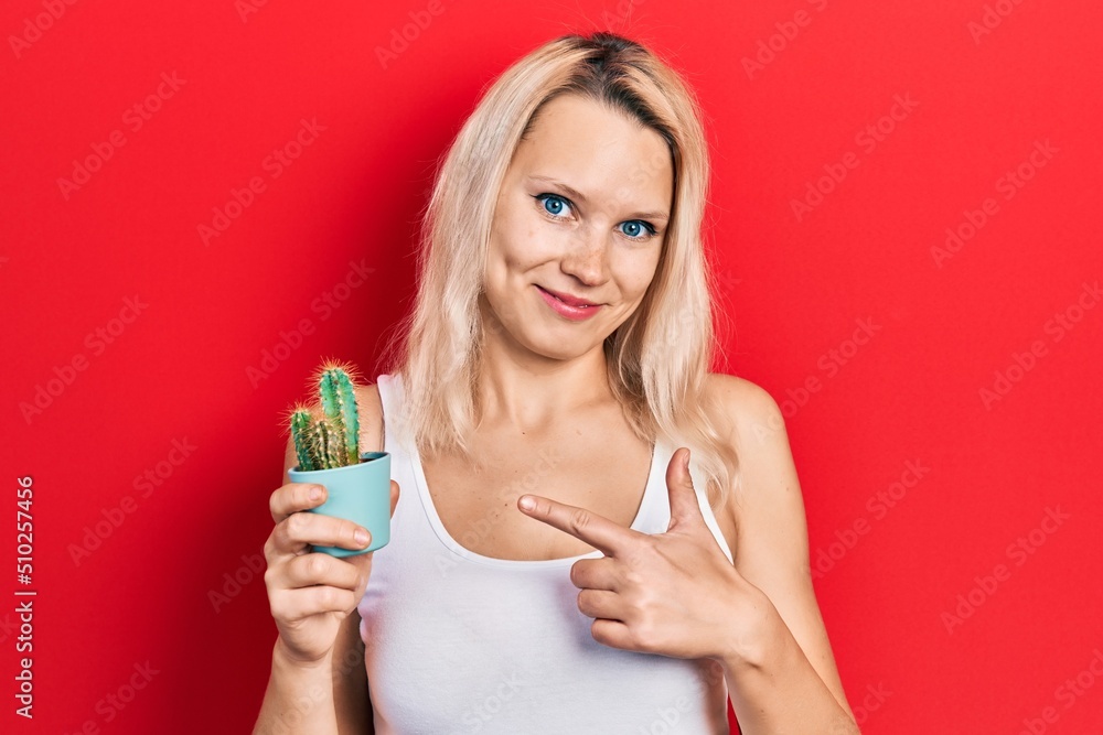 Beautiful caucasian blonde woman holding small cactus pot smiling happy pointing with hand and finger Beautiful caucasian blonde woman holding small cactus pot smiling happy pointing with hand and finger
