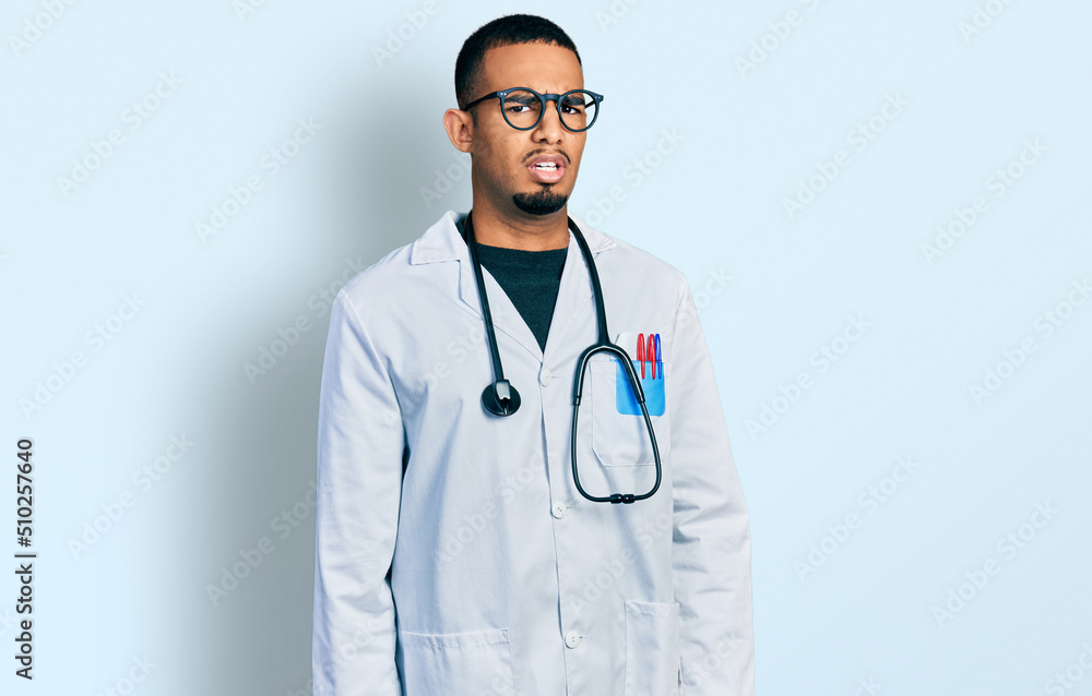 Young african american man wearing doctor uniform and stethoscope in shock face, looking skeptical and sarcastic, surprised with open mouth