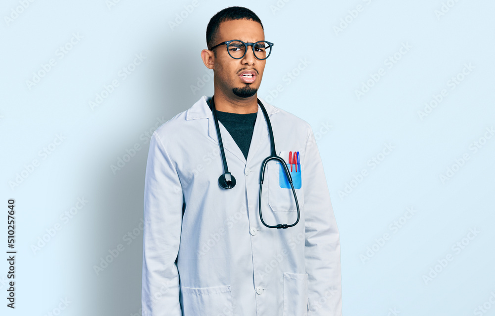 Young african american man wearing doctor uniform and stethoscope in shock face, looking skeptical and sarcastic, surprised with open mouth Young african american man wearing doctor uniform and stethoscope in shock face, looking skeptical and sarcastic, surprised with open mouth