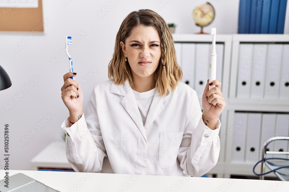 Young hispanic dentist woman holding electric toothbrush and teethbrush at clinic clueless and confused expression. doubt concept. Young hispanic dentist woman holding electric toothbrush and teethbrush at clinic clueless and confused expression. doubt concept.