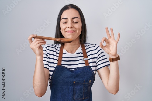 Tablou pe pânză Young brunette woman wearing apron tasting food holding wooden spoon doing ok si