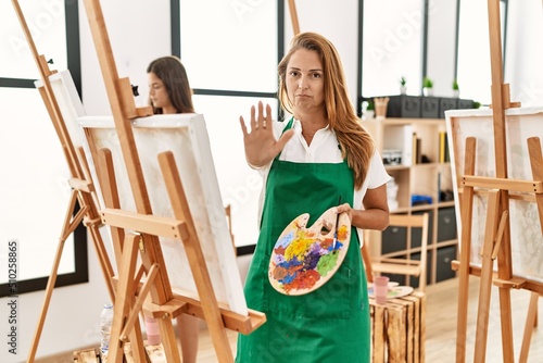 Young hispanic middle age woman at art classroom with open hand doing stop sign with serious and confident expression, defense gesture