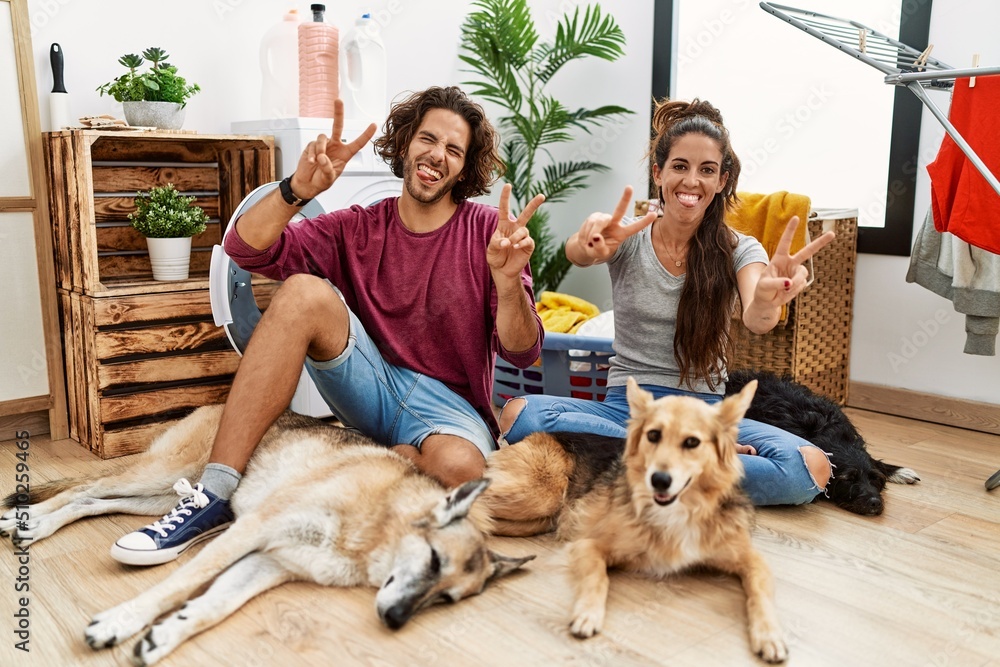 Young hispanic couple doing laundry with dogs smiling with tongue out showing fingers of both hands doing victory sign. number two.