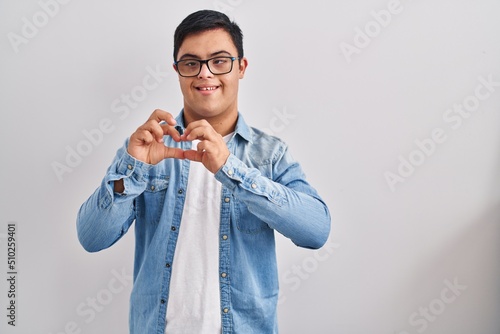 Young hispanic man with down syndrome wearing casual denim jacket over white background smiling in love doing heart symbol shape with hands. romantic concept.