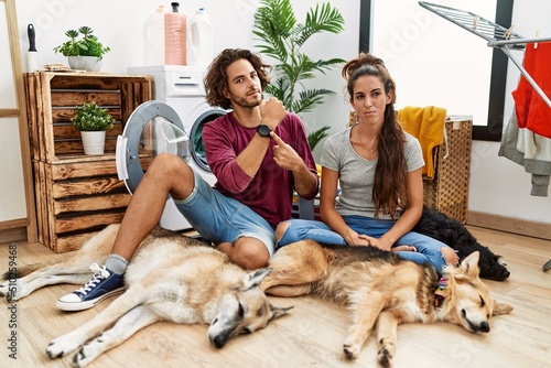Photography Young hispanic couple doing laundry with dogs in hurry pointing to watch time, i