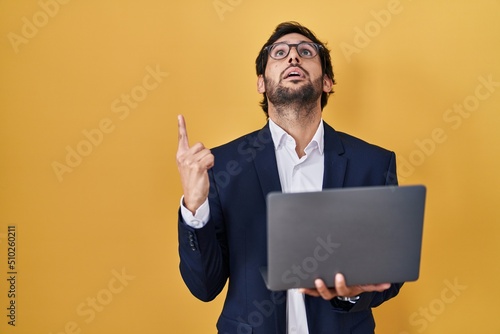 Handsome latin man working using computer laptop amazed and surprised looking up and pointing with fingers and raised arms.
