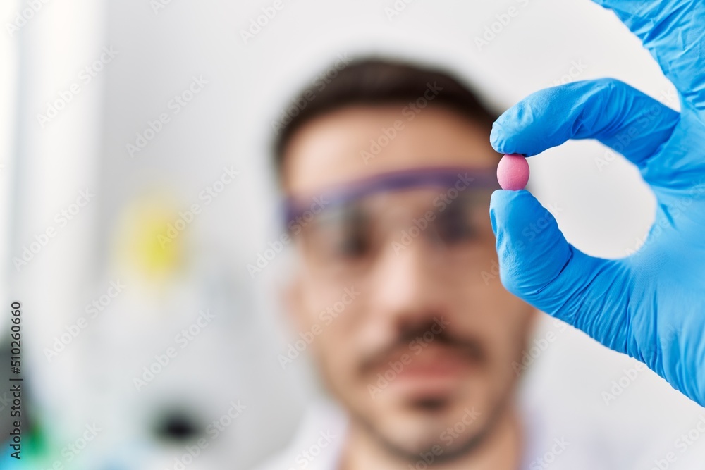 Naklejka premium Young hispanic man wearing scientist uniform holding pill at laboratory