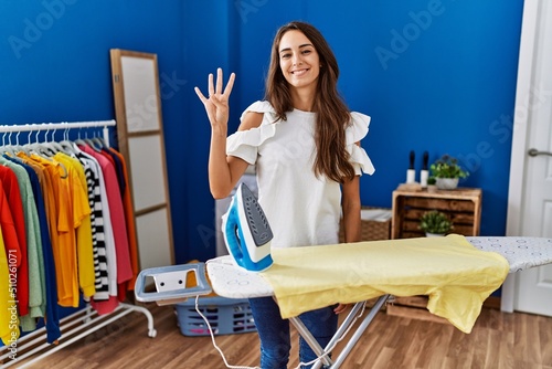 Young hispanic woman ironing clothes at laundry room showing and pointing up with fingers number four while smiling confident and happy.