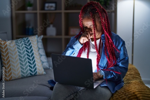 African american woman with braided hair using computer laptop at night smelling something stinky and disgusting, intolerable smell, holding breath with fingers on nose. bad smell