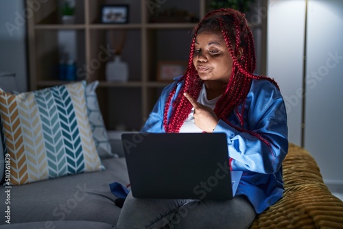 African american woman with braided hair using computer laptop at night pointing with hand finger to the side showing advertisement, serious and calm face