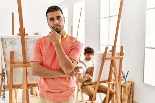 Young hispanic man at art studio looking confident at the camera smiling with crossed arms and hand raised on chin. thinking positive.