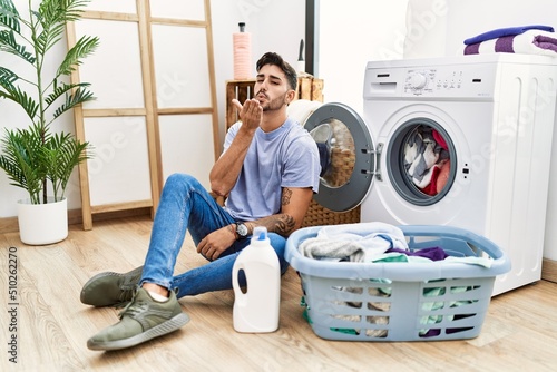 Young hispanic man putting dirty laundry into washing machine looking at the camera blowing a kiss with hand on air being lovely and sexy. love expression.