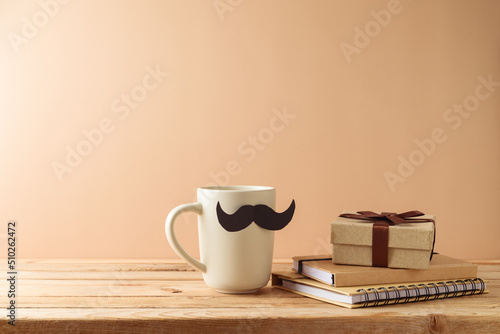 Father's day concept with coffee cup, mustache, notebook and gift box on wooden table over beige background