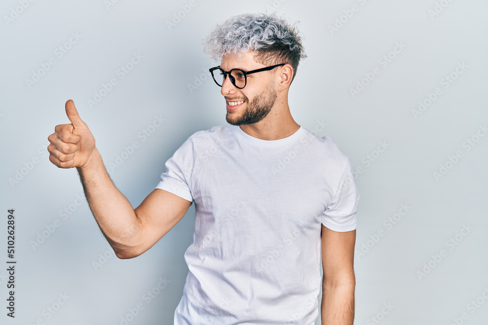 Young hispanic man with modern dyed hair wearing white t shirt and glasses looking proud, smiling doing thumbs up gesture to the side Young hispanic man with modern dyed hair wearing white t shirt and glasses looking proud, smiling doing thumbs up gesture to the side