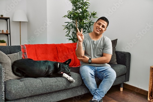 Young latin man and dog sitting on the sofa at home smiling looking to the camera showing fingers doing victory sign. number two. © Krakenimages.com