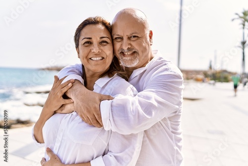 Middle age hispanic couple of husband and wife together walking by the beach on a sunny day. Hugging in love on vacation to the seaside.