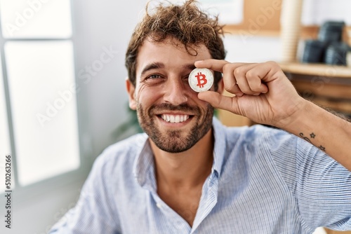 Young hispanic businessman smiling happy holding bitcoin over eye at the office. photo