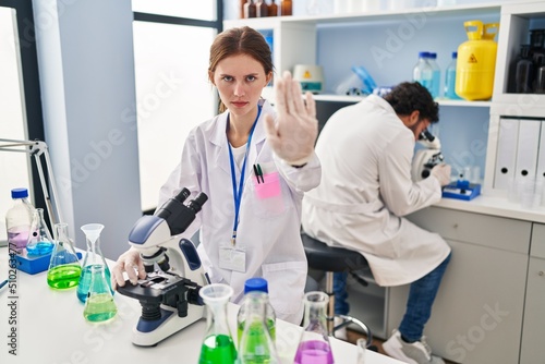 Young two people working at scientist laboratory with open hand doing stop sign with serious and confident expression  defense gesture