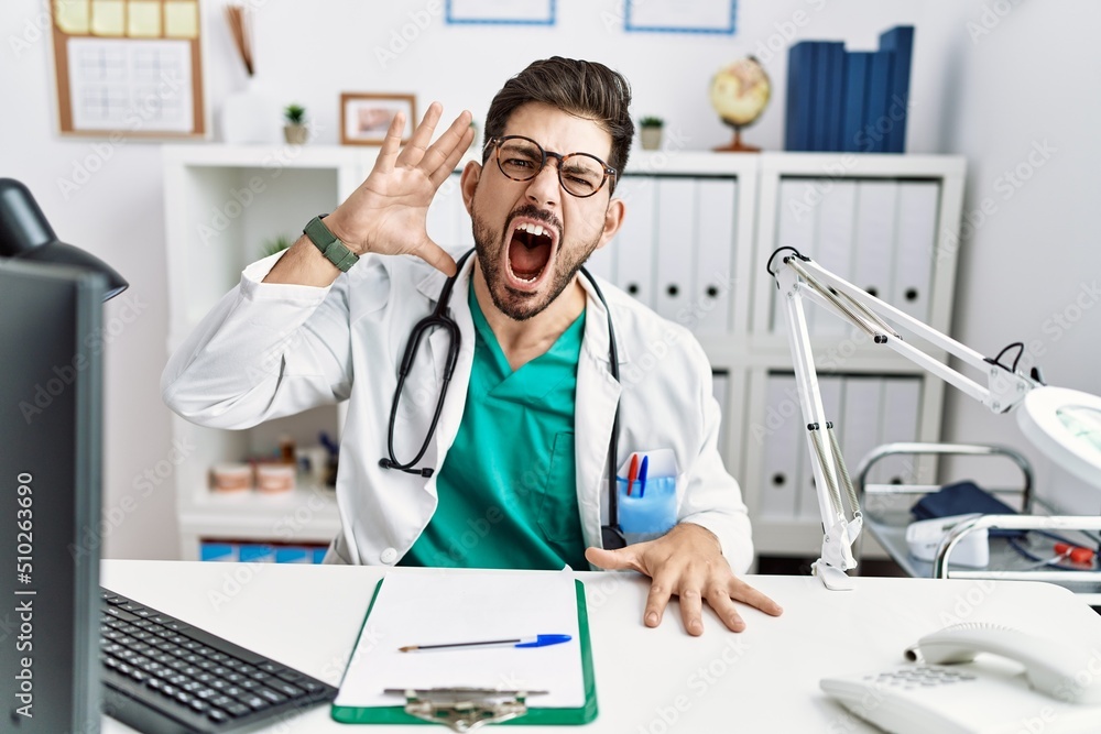 Young man with beard wearing doctor uniform and stethoscope at the clinic shouting and screaming loud to side with hand on mouth. communication concept.