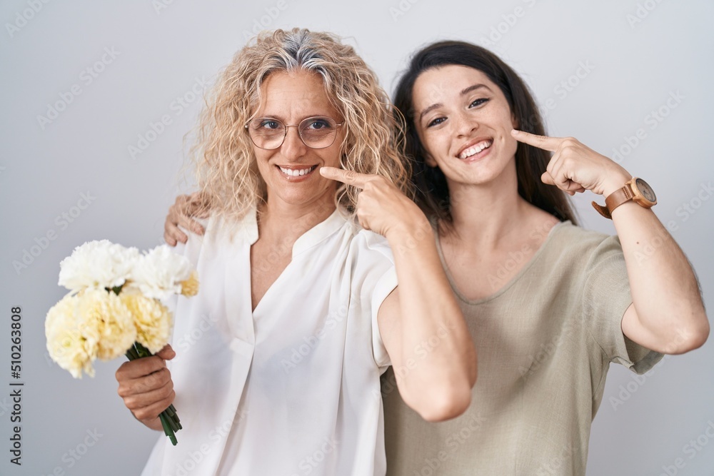 Mother and daughter holding bouquet of white flowers smiling cheerful showing and pointing with fingers teeth and mouth. dental health concept. Mother and daughter holding bouquet of white flowers smiling cheerful showing and pointing with fingers teeth and mouth. dental health concept.