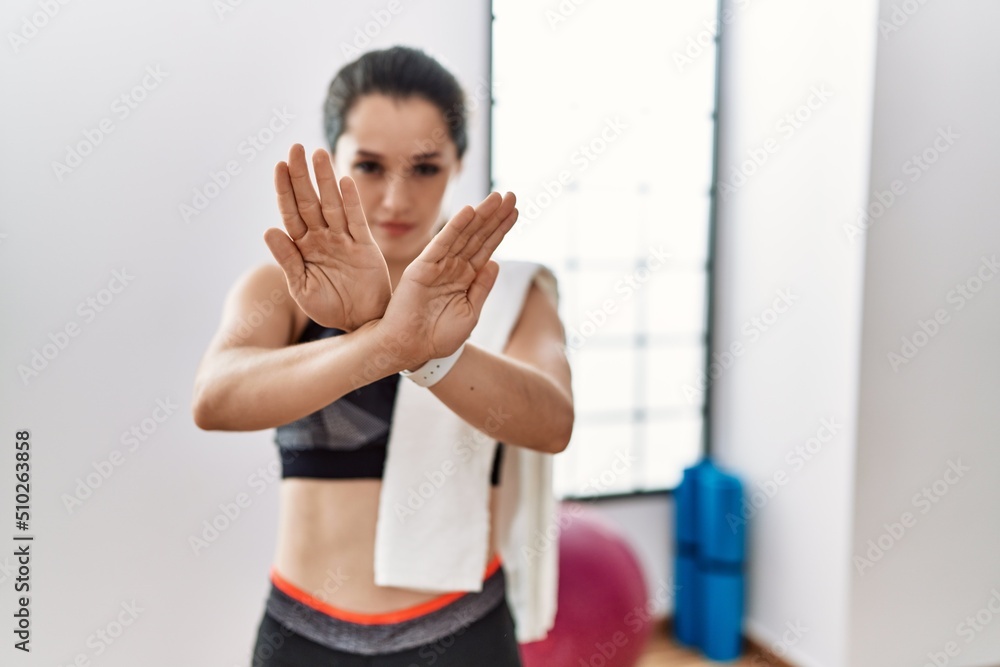 Young brunette woman wearing sportswear and towel at the gym rejection expression crossing arms and palms doing negative sign, angry face