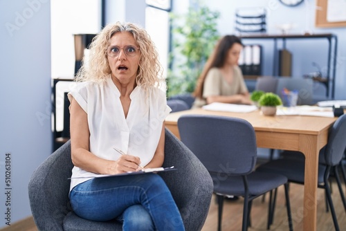 Middle age woman working at the office holding clipboard scared and amazed with open mouth for surprise, disbelief face