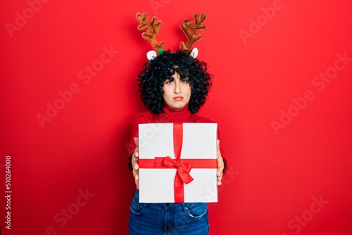 Young middle east woman wearing deer christmas hat holding gift relaxed with serious expression on face. simple and natural looking at the camera.