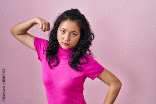 Young asian woman standing over pink background strong person showing arm muscle, confident and proud of power