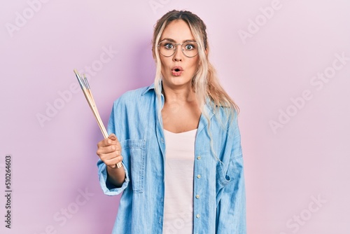 Beautiful young blonde woman holding paintbrushes scared and amazed with open mouth for surprise  disbelief face