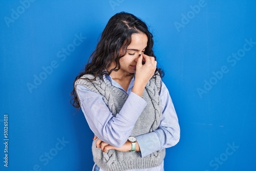 Young brunette woman standing over blue background tired rubbing nose and eyes feeling fatigue and headache. stress and frustration concept. © Krakenimages.com