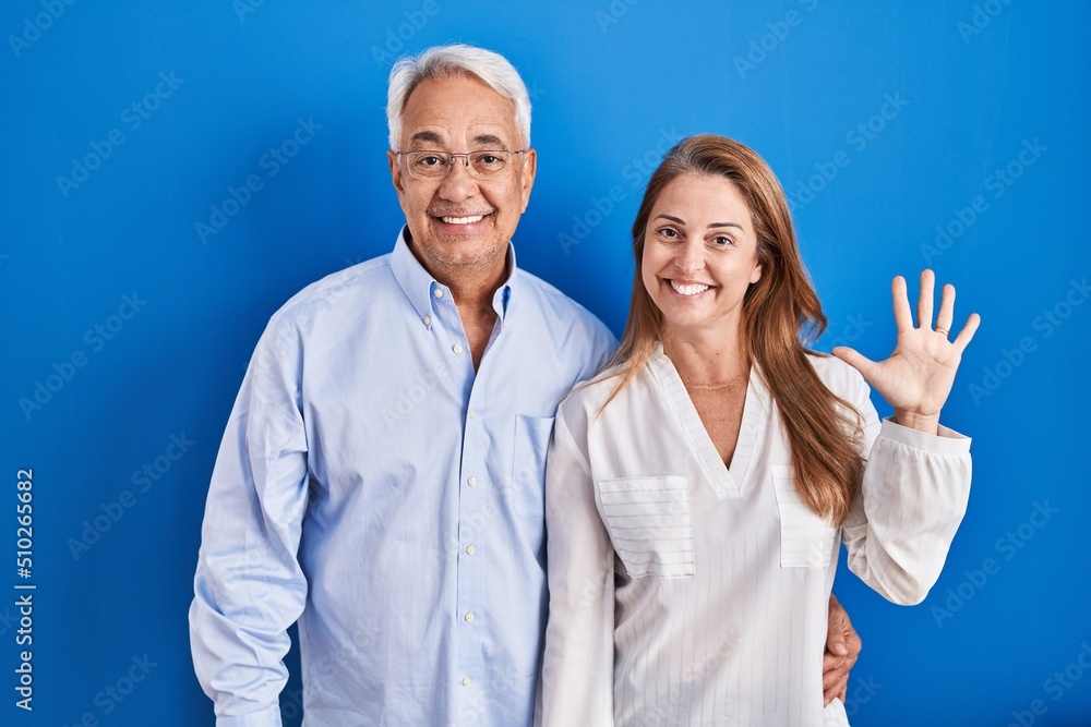 Middle age hispanic couple standing over blue background showing and pointing up with fingers number five while smiling confident and happy. Middle age hispanic couple standing over blue background showing and pointing up with fingers number five while smiling confident and happy.