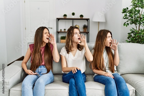 Group of three hispanic girls sitting on the sofa at home shouting and screaming loud to side with hand on mouth. communication concept.