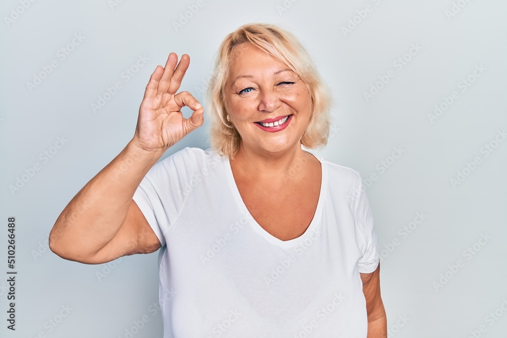 Middle age blonde woman doing ok positive gesture winking looking at the camera with sexy expression, cheerful and happy face. Middle age blonde woman doing ok positive gesture winking looking at the camera with sexy expression, cheerful and happy face.