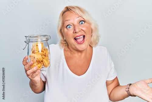 Middle age blonde woman holding uncooked noodles celebrating achievement with happy smile and winner expression with raised hand