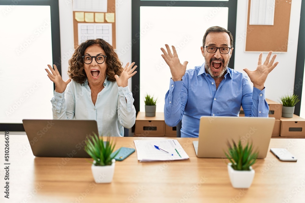 Middle age hispanic woman and man sitting with laptop at the office celebrating crazy and amazed for success with arms raised and open eyes screaming excited. winner concept Middle age hispanic woman and man sitting with laptop at the office celebrating crazy and amazed for success with arms raised and open eyes screaming excited. winner concept