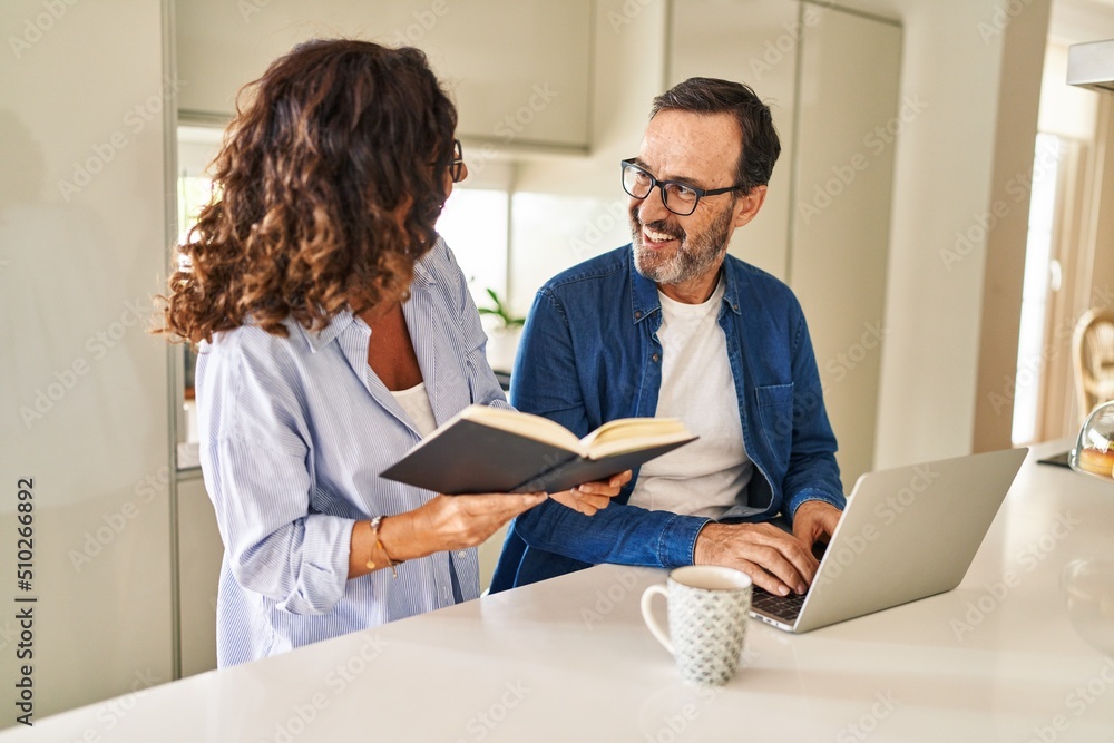 Middle age hispanic couple reading book and using laptop at kitchen Middle age hispanic couple reading book and using laptop at kitchen