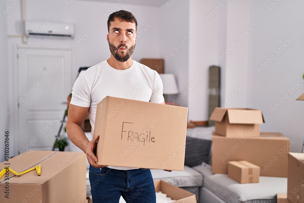 Handsome hispanic man moving to a new home holding fragile box making fish face with mouth and squinting eyes, crazy and comical. Handsome hispanic man moving to a new home holding fragile box making fish face with mouth and squinting eyes, crazy and comical.