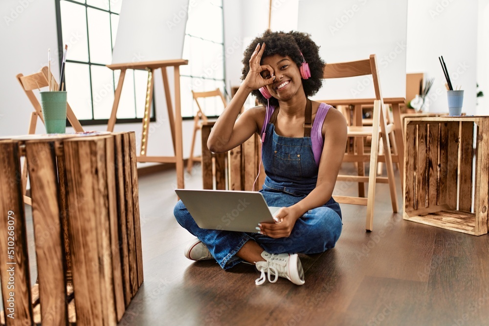 Young african american woman with afro hair at art studio using laptop smiling happy doing ok sign with hand on eye looking through fingers Young african american woman with afro hair at art studio using laptop smiling happy doing ok sign with hand on eye looking through fingers