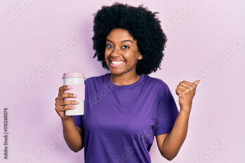 Young african american woman holding coffee pointing thumb up to the side smiling happy with open mouth