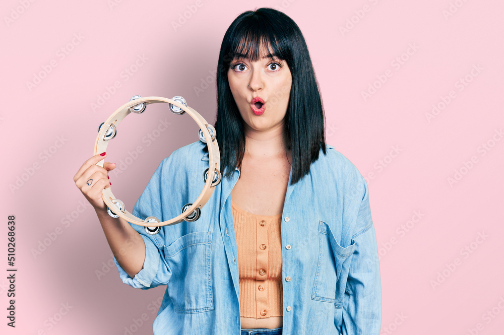 Young hispanic plus size woman playing tambourine scared and amazed with open mouth for surprise, disbelief face