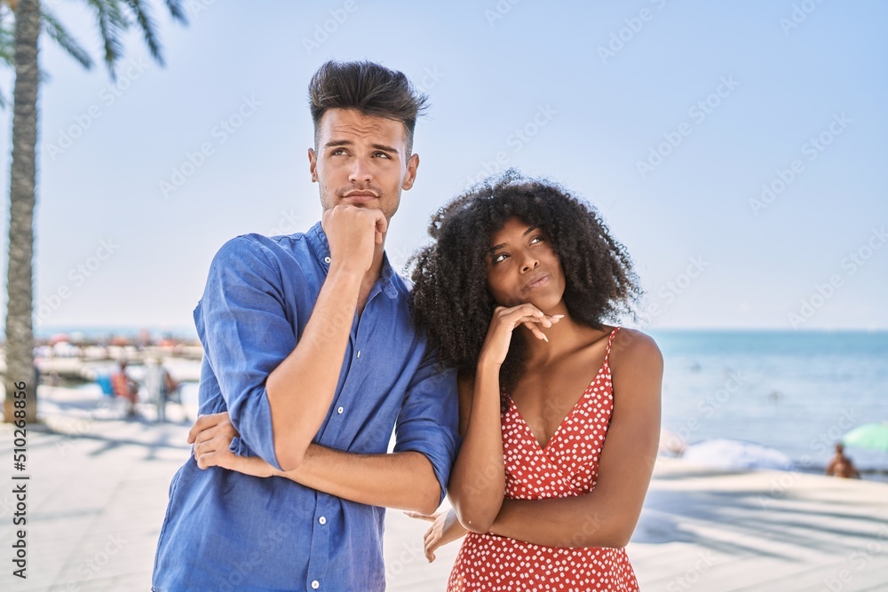 Young interracial couple outdoors on a sunny day with hand on chin thinking about question, pensive expression. smiling with thoughtful face. doubt concept.