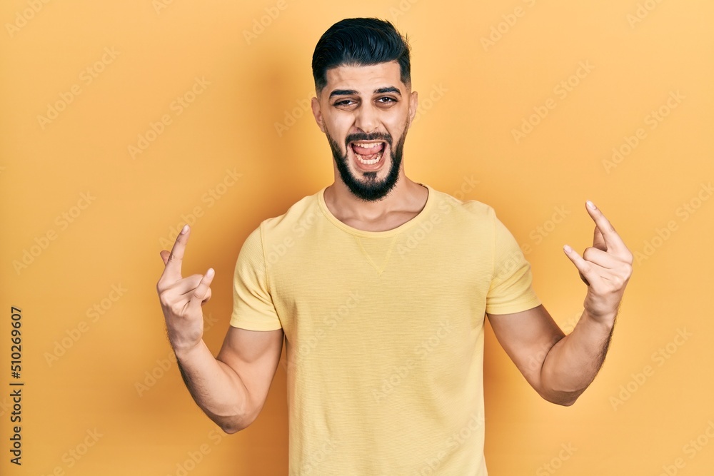 Handsome man with beard wearing casual yellow t shirt shouting with crazy expression doing rock symbol with hands up. music star. heavy concept.