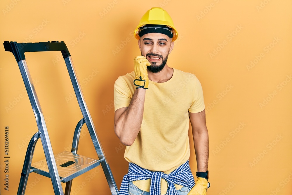 Handsome man with beard by construction stairs wearing hardhat looking stressed and nervous with hands on mouth biting nails. anxiety problem. Handsome man with beard by construction stairs wearing hardhat looking stressed and nervous with hands on mouth biting nails. anxiety problem.