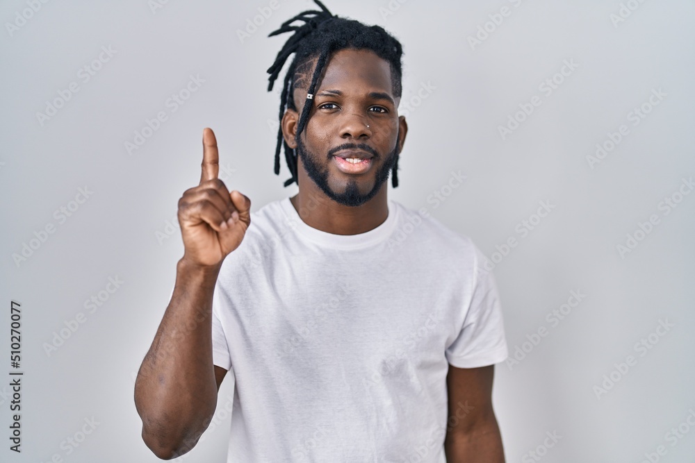 African man with dreadlocks wearing casual t shirt over white background showing and pointing up with finger number one while smiling confident and happy. African man with dreadlocks wearing casual t shirt over white background showing and pointing up with finger number one while smiling confident and happy.