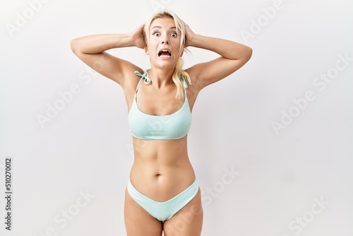 Young caucasian woman wearing bikini over isolated background crazy and scared with hands on head, afraid and surprised of shock with open mouth