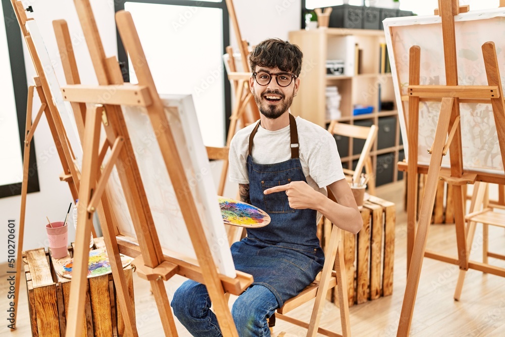 Hispanic man with beard at art studio smiling happy pointing with hand and finger Hispanic man with beard at art studio smiling happy pointing with hand and finger